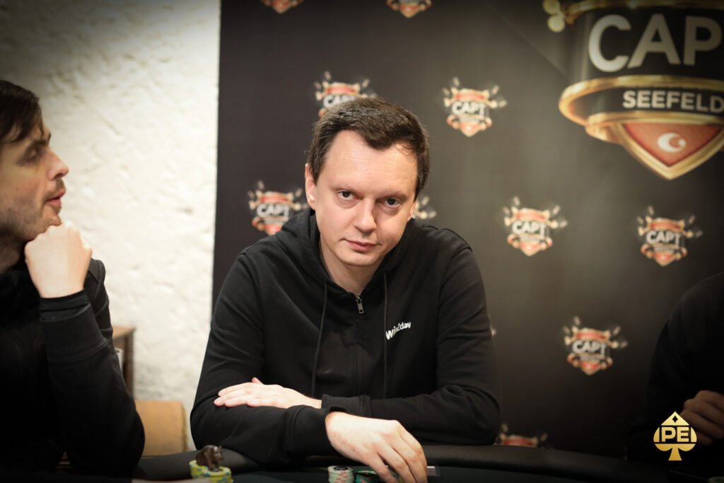 Ein Mann in einem schwarzen Anzug und einer roten Krawatte sitzt an einem Pokertisch und hält seine Karten in der Hand. Er schaut konzentriert auf den Tisch und wirkt entschlossen.