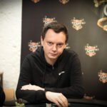 Ein Mann in einem schwarzen Anzug und einer roten Krawatte sitzt an einem Pokertisch und hält seine Karten in der Hand. Er schaut konzentriert auf den Tisch und wirkt entschlossen.
