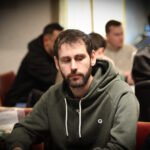 Ein Mann in einem Anzug sitzt an einem Pokertisch und hält eine Hand mit Spielkarten. Um ihn herum stehen andere Spieler und beobachten das Spiel.