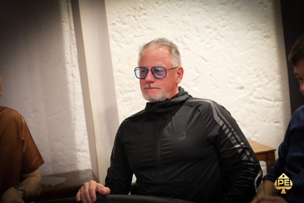 Ein Mann in einem dunkelblauen Anzug sitzt an einem Pokertisch und hält seine Karten. Er schaut konzentriert auf den Tisch, umgeben von anderen Spielern und Pokerchips.