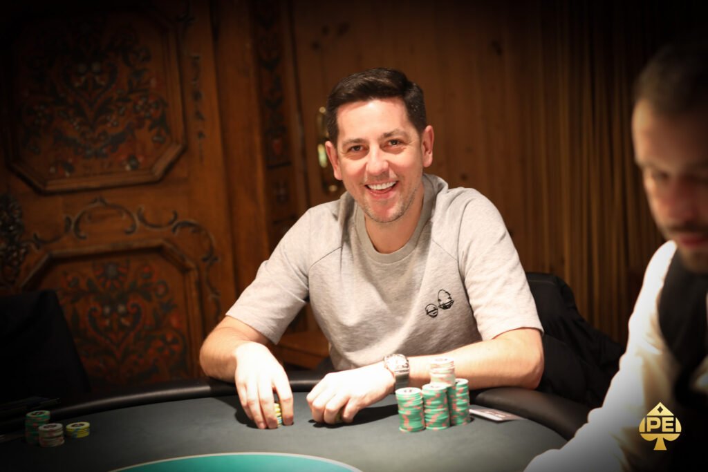 Ein Mann in einem dunklen Anzug und einer Krawatte sitzt an einem Pokertisch und betrachtet konzentriert seine Karten. Im Hintergrund sind weitere Spieler und Pokerchips zu sehen.