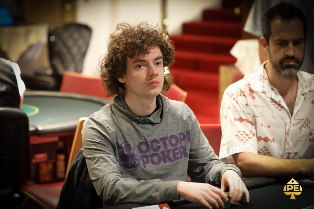 Ein lustiger Moment bei einem Pokerturnier: Zwei Spieler lachen und stoßen mit ihren Chips aneinander, während sie ihre Karten betrachten. Im Hintergrund sitzen weitere Spieler am Tisch.