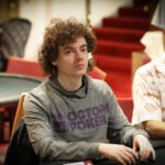 Ein lustiger Moment bei einem Pokerturnier: Zwei Spieler lachen und stoßen mit ihren Chips aneinander, während sie ihre Karten betrachten. Im Hintergrund sitzen weitere Spieler am Tisch.