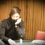 Ein Mann und eine Frau sitzen lächelnd an einem Pokertisch, während sie Karten halten und Chips stapeln. Die Frau hat blonde Haare und trägt ein rotes Oberteil, der Mann hat dunkle Haare