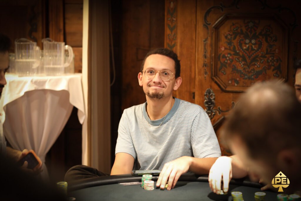 Ein professioneller Pokerspieler sitzt an einem Spieltisch und betrachtet konzentriert seine Karten. Im Hintergrund sind weitere Spieler und Pokerchips zu sehen.