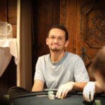 Ein professioneller Pokerspieler sitzt an einem Spieltisch und betrachtet konzentriert seine Karten. Im Hintergrund sind weitere Spieler und Pokerchips zu sehen.