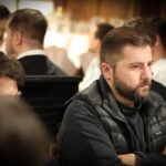 Ein Mann in einem dunkelblauen Anzug sitzt lächelnd an einem Pokertisch und hält zwei Spielkarten in der Hand. Im Hintergrund sind Pokerchips und weitere Spieler zu sehen.