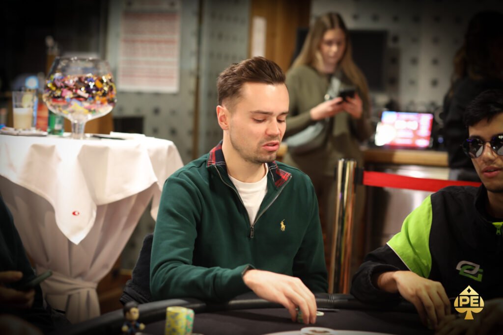 Ein Mann sitzt an einem Pokertisch mit grüner Filzdecke und hält in seiner Hand zwei Spielkarten. Er trägt einen dunklen Anzug und eine schwarze Krawatte. Im Hintergrund sind Spiel