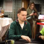 Ein Mann sitzt an einem Pokertisch mit grüner Filzdecke und hält in seiner Hand zwei Spielkarten. Er trägt einen dunklen Anzug und eine schwarze Krawatte. Im Hintergrund sind Spiel