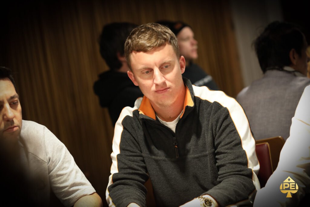 Ein Mann in einem eleganten Anzug sitzt an einem Pokertisch und hält Pokerchips in der Hand, während er ernsthaft auf seine Karten schaut. Im Hintergrund sind weitere Spieler und ein Stapel Pokerch