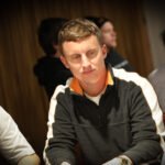 Ein Mann in einem eleganten Anzug sitzt an einem Pokertisch und hält Pokerchips in der Hand, während er ernsthaft auf seine Karten schaut. Im Hintergrund sind weitere Spieler und ein Stapel Pokerch