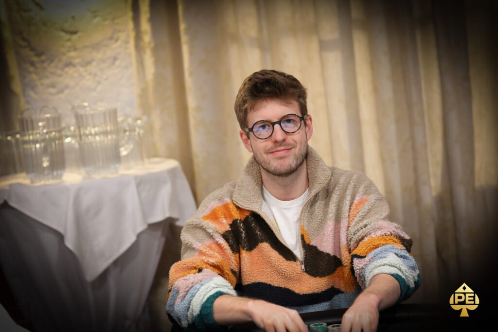 Ein Mann in einem schwarzen Anzug sitzt an einem Pokertisch und hält seine Spielkarten in der Hand. Im Hintergrund sind weitere Spieler und Chips zu sehen.