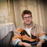 Ein Mann in einem schwarzen Anzug sitzt an einem Pokertisch und hält seine Spielkarten in der Hand. Im Hintergrund sind weitere Spieler und Chips zu sehen.