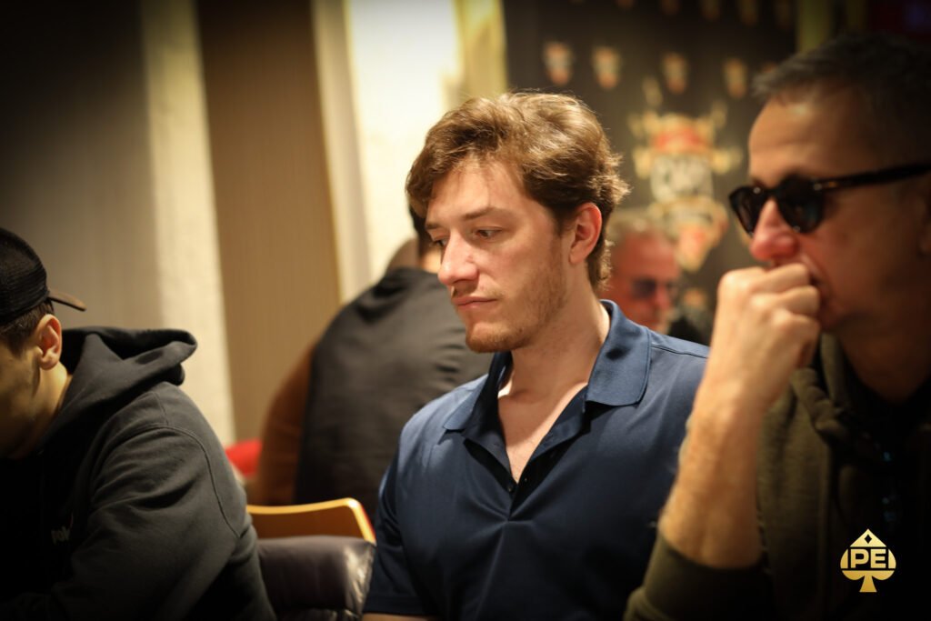 Ein farbenfrohes Bild von einem Pokertisch mit grünen Filz und Chips, Karten und Getränken darauf. In der Mitte sitzen vier Personen und spielen Poker.