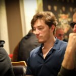 Ein farbenfrohes Bild von einem Pokertisch mit grünen Filz und Chips, Karten und Getränken darauf. In der Mitte sitzen vier Personen und spielen Poker.