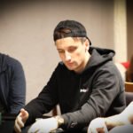 Ein Mann in einem dunkelblauen Anzug und Krawatte sitzt an einem Pokertisch und hält seine Spielkarten in der Hand. Er schaut konzentriert auf den Tisch, umgeben von Pokerchips