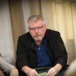 Ein Mann spielt Poker an einem Tisch in einem eleganten Casino. Er hält Karten in der Hand und hat einen ernsten Ausdruck im Gesicht, während andere Spieler im Hintergrund zu sehen sind.
