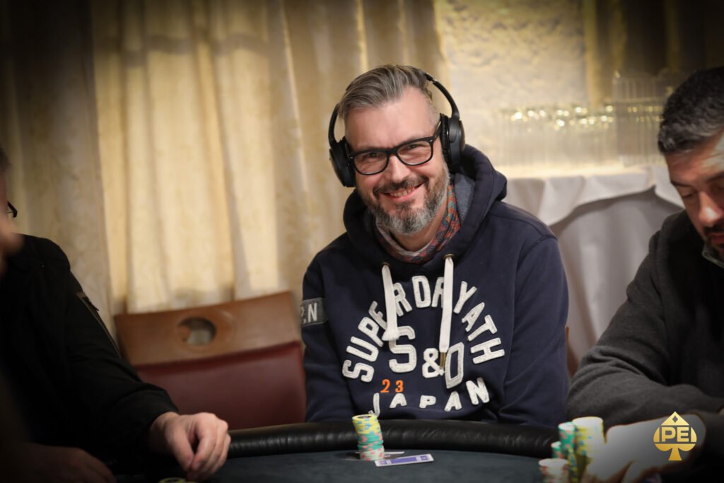 Ein Mann sitzt an einem Pokertisch und hält Pokerchips in der Hand. Er trägt einen dunklen Anzug und eine Sonnenbrille. Andere Spieler und Karten sind im Hintergrund zu sehen.