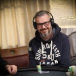 Ein Mann sitzt an einem Pokertisch und hält Pokerchips in der Hand. Er trägt einen dunklen Anzug und eine Sonnenbrille. Andere Spieler und Karten sind im Hintergrund zu sehen.