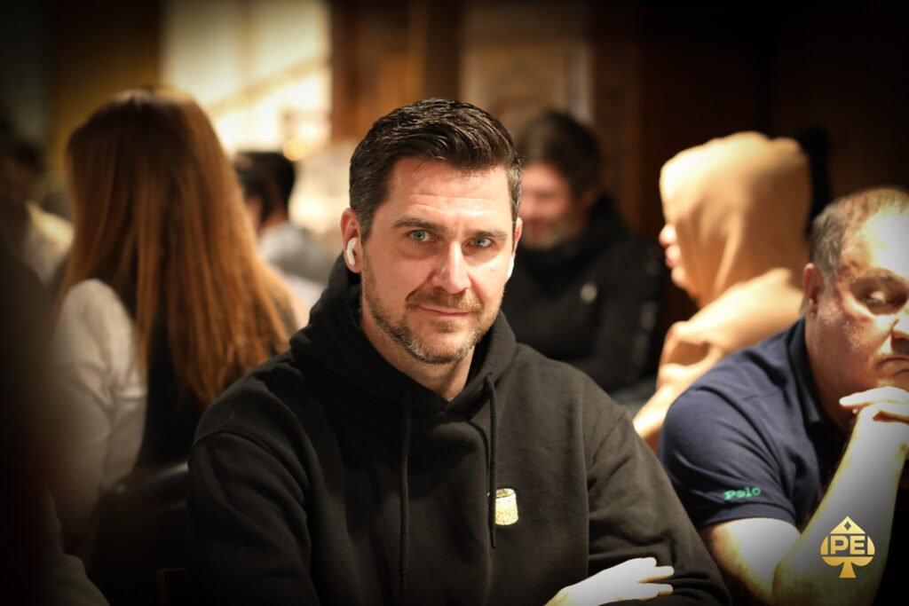 Ein Mann und eine Frau sitzen an einem Pokertisch und betrachten ihre Karten. Der Mann hat einen Stapel Chips vor sich liegen, während die Frau konzentriert auf ihre Hand schaut. Im Hintergrund sind weitere