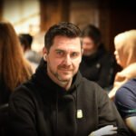 Ein Mann und eine Frau sitzen an einem Pokertisch und betrachten ihre Karten. Der Mann hat einen Stapel Chips vor sich liegen, während die Frau konzentriert auf ihre Hand schaut. Im Hintergrund sind weitere