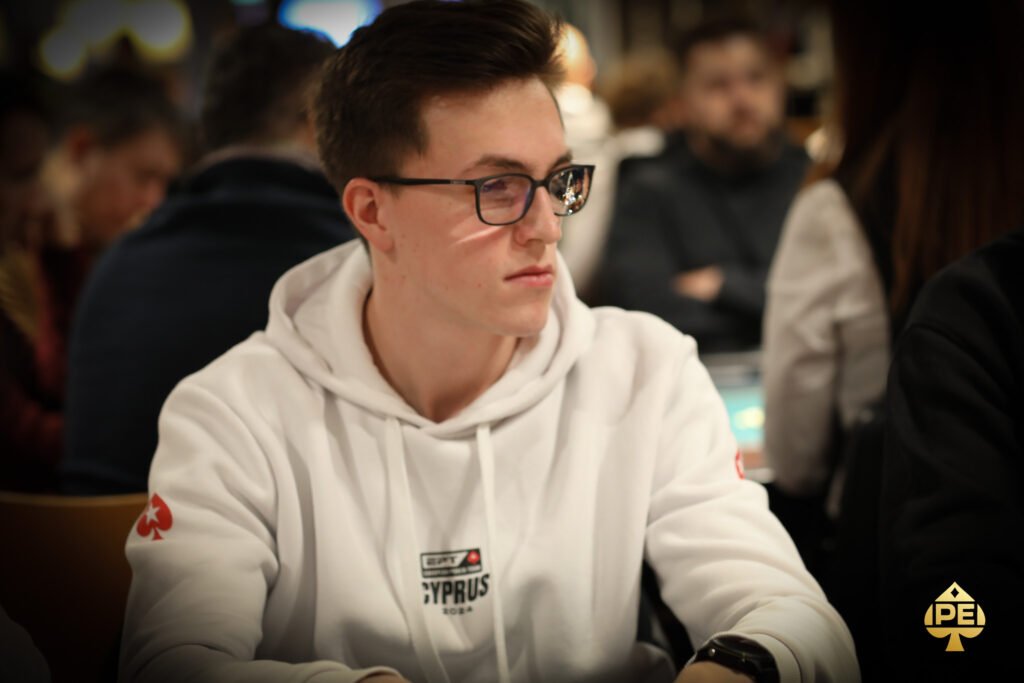 Ein Mann in einem schwarzen Anzug und einer Krawatte sitzt an einem Pokertisch und hält seine Karten in der Hand, während er konzentriert auf seine Gegner schaut.