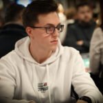 Ein Mann in einem schwarzen Anzug und einer Krawatte sitzt an einem Pokertisch und hält seine Karten in der Hand, während er konzentriert auf seine Gegner schaut.