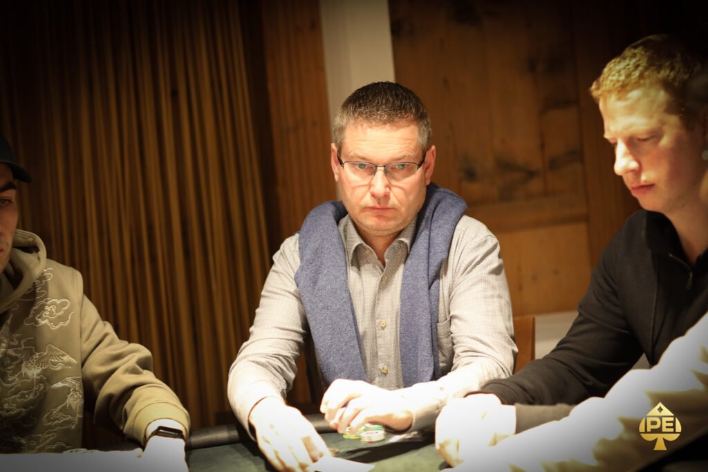 Ein Mann mit einer schwarzen Kappe und einem schwarzen Anzug, der an einem Pokertisch sitzt und Karten hält, während er konzentriert in die Kamera schaut. Im Hintergrund sind weitere Pokers