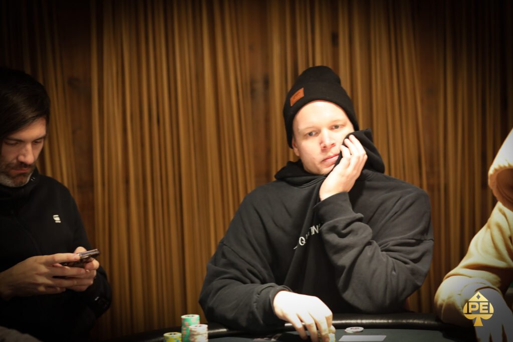 Ein Mann und eine Frau sitzen in einem eleganten Casino und spielen Poker. Der Mann trägt einen Anzug und eine Krawatte, die Frau hat ein elegantes Abendkleid an. Sie haben ernste Gesichter und halten