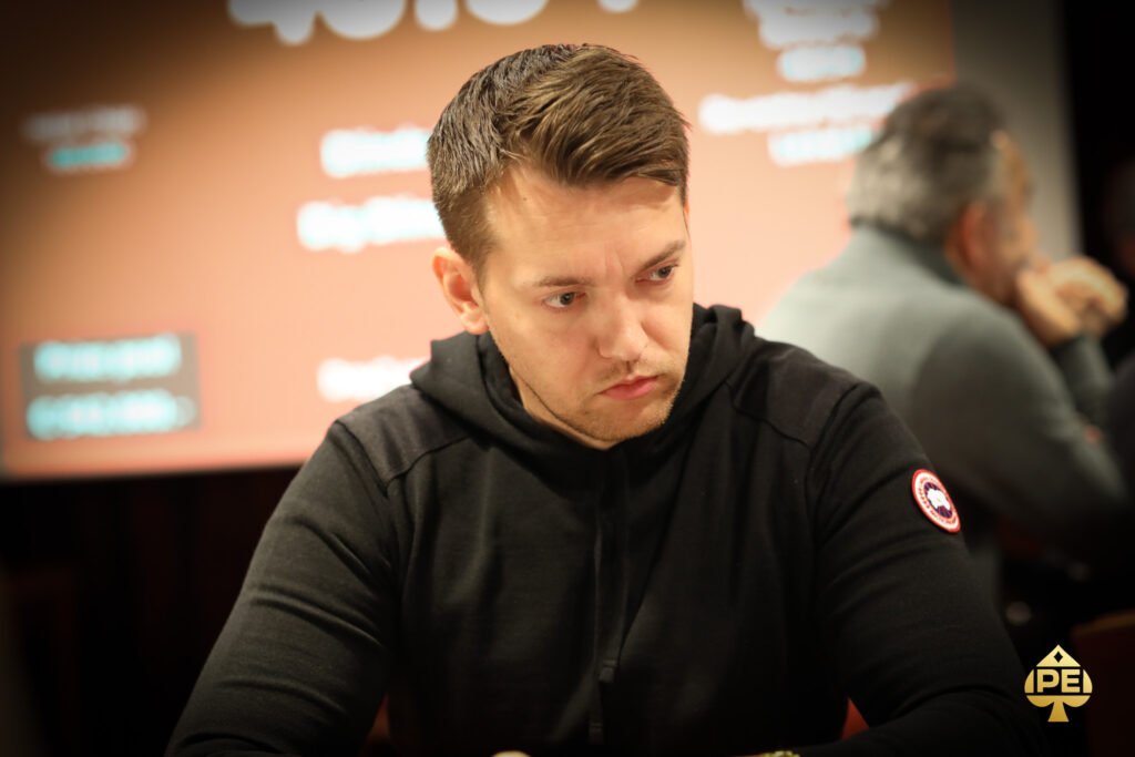 Ein Mann sitzt an einem Pokertisch mit grünen Filz und hält zwei Asse in der Hand. Er hat eine ernste Miene und trägt einen dunklen Anzug. Im Hintergrund sind weitere Spieler und Chips