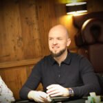 Ein Mann und eine Frau sitzen an einem Pokertisch und spielen Karten. Der Mann hat einen Stapel Chips vor sich und sieht konzentriert aus, während die Frau ihre Karten betrachtet. Im Hintergrund sind weitere