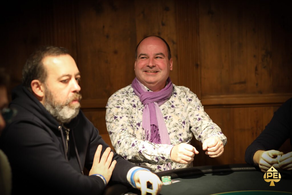 Ein professioneller Pokerspieler mit Sonnenbrille und Kappe sitzt an einem Pokertisch mit grüner Filzdecke. Er betrachtet konzentriert seine Karten während er in einer entspannten H