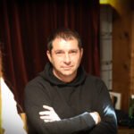 Ein Mann sitzt an einem Pokertisch in einem eleganten Casino. Er trägt einen schwarzen Anzug und hat eine ernste Miene, während er seine Karten betrachtet. Im Hintergrund sind andere Spieler und Dealer zu