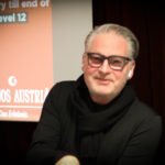 Ein Mann sitzt an einem Pokertisch mit grünem Filz. Er hält zwei Spielkarten in der Hand und schaut konzentriert auf den Tisch. Um ihn herum sind Pokerchips und Spielkarten