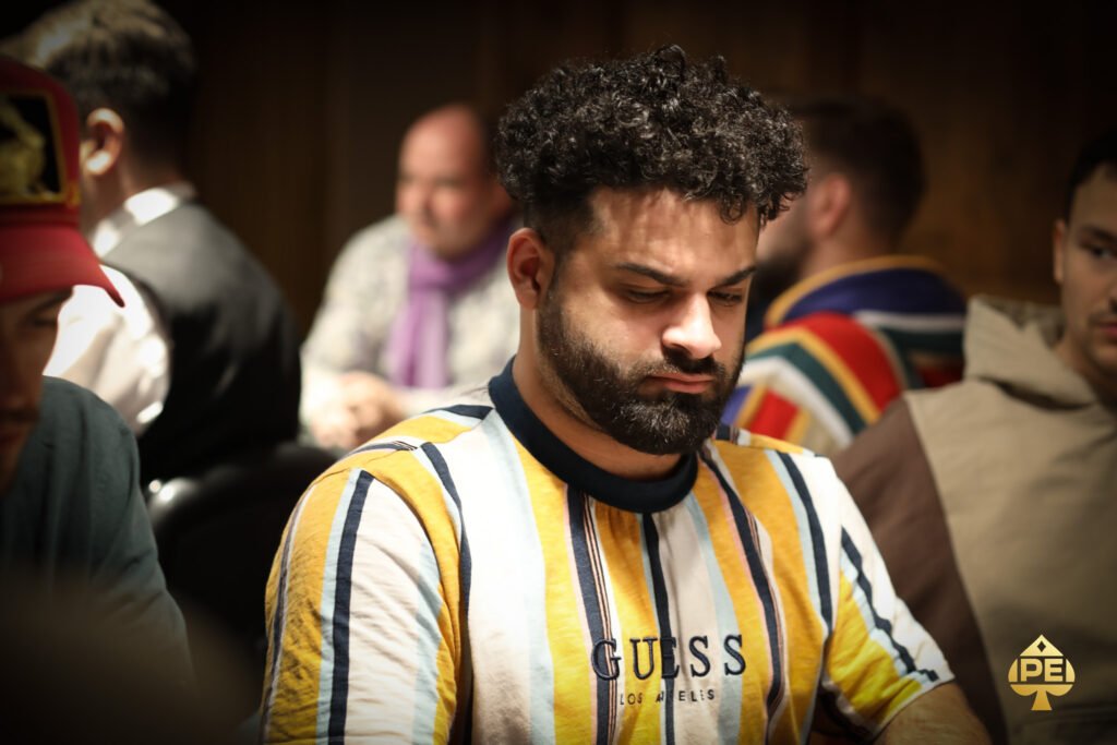 Ein Mann und eine Frau sitzen an einem Pokertisch und schauen konzentriert auf ihre Karten. Der Mann trägt einen dunklen Anzug, die Frau ein elegantes Kleid. Im Hintergrund sind Dealer und andere