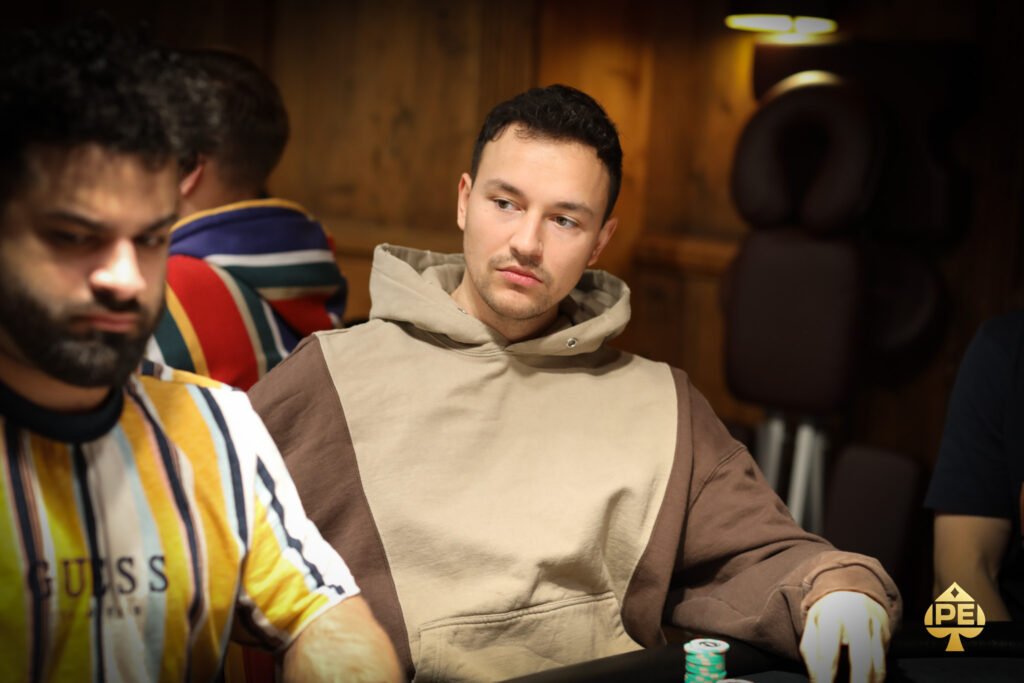 Ein Mann in einem dunklen Anzug sitzt an einem Pokertisch und hält zwei Spielkarten in der Hand. Er wirkt konzentriert und studiert seine Karten. Im Hintergrund sind weitere Spieler und Spielch