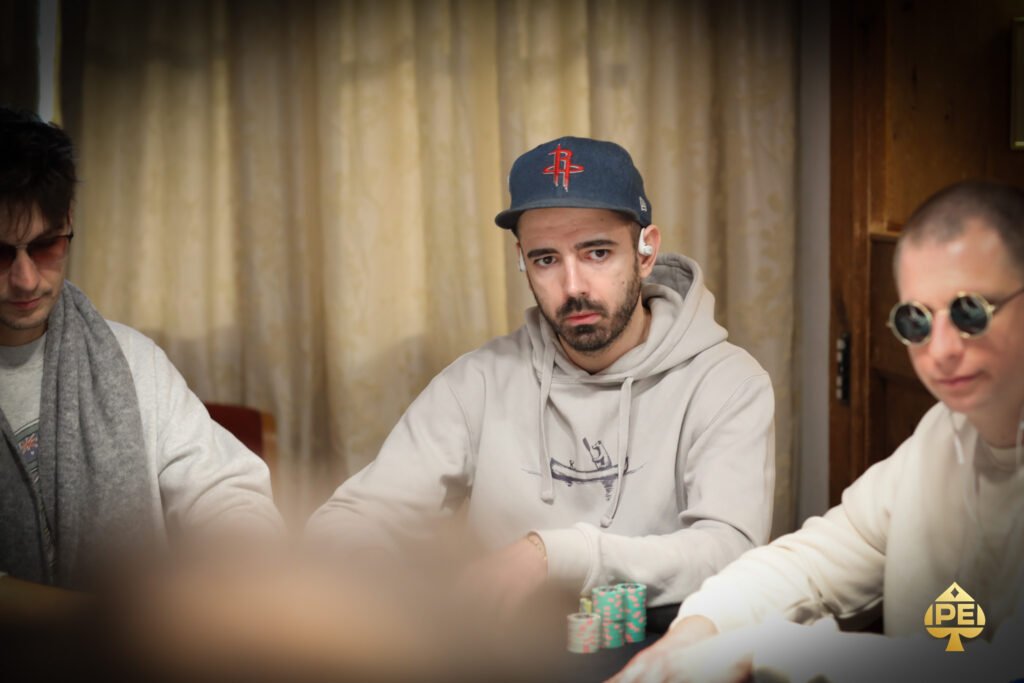 Ein Mann sitzt an einem Pokertisch, hält Karten in der Hand und betrachtet konzentriert seine Mitspieler. Im Hintergrund sind Pokerchips und Getränke zu sehen.