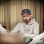 Ein Mann sitzt an einem Pokertisch, hält Karten in der Hand und betrachtet konzentriert seine Mitspieler. Im Hintergrund sind Pokerchips und Getränke zu sehen.
