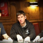 Ein Mann sitzt an einem Pokertisch und hält zwei Karten in der Hand, während er konzentriert auf die Gemeinschaftskarten auf dem Tisch schaut. Im Hintergrund sind weitere Spieler zu sehen.