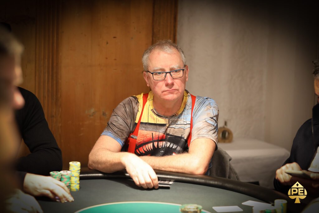 Ein Mann in einem eleganten Anzug und einer Sonnenbrille sitzt an einem Pokertisch und hält seine Karten in der Hand, während er mit ernstem Gesichtsausdruck auf seine Gegner schaut.