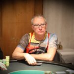 Ein Mann in einem eleganten Anzug und einer Sonnenbrille sitzt an einem Pokertisch und hält seine Karten in der Hand, während er mit ernstem Gesichtsausdruck auf seine Gegner schaut.