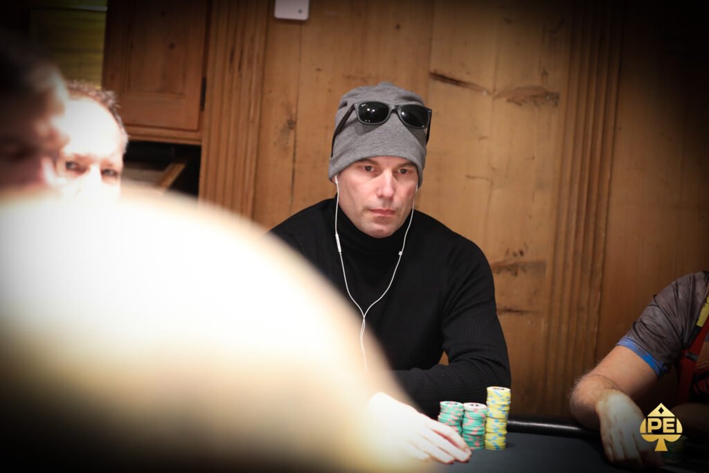 Ein glücklicher Mann in einem schwarzen Anzug und einer Fliege, der lächelnd eine Handkarte mit Pik-As und Pik-König hält, während er an einem Pokertisch sitzt. Im H