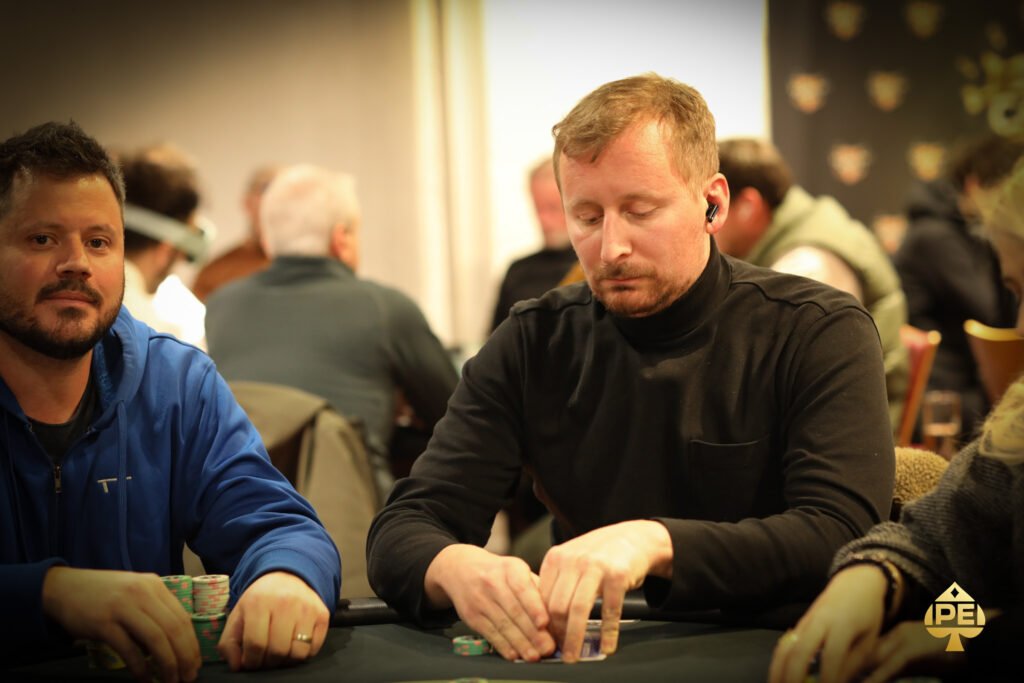 Ein junger Mann sitzt an einem Pokertisch und hält seine Karten in der Hand. Er hat einen konzentrierten Ausdruck im Gesicht und schaut auf den Tisch. Im Hintergrund sind andere Spieler und