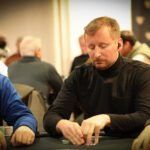 Ein junger Mann sitzt an einem Pokertisch und hält seine Karten in der Hand. Er hat einen konzentrierten Ausdruck im Gesicht und schaut auf den Tisch. Im Hintergrund sind andere Spieler und