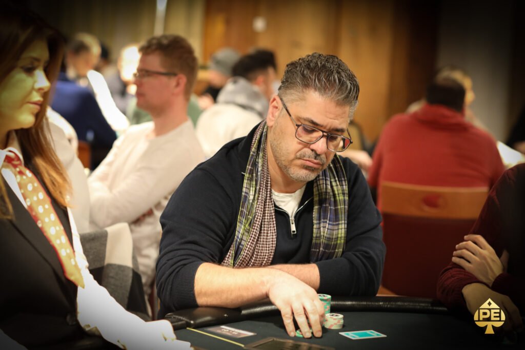 Ein professioneller Pokerspieler sitzt an einem Tisch und hält seine Karten in der Hand. Er wirkt konzentriert und fokussiert, während er an einem Turnier teilnimmt. Die Umgebung ist