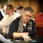 Ein professioneller Pokerspieler sitzt an einem Tisch und hält seine Karten in der Hand. Er wirkt konzentriert und fokussiert, während er an einem Turnier teilnimmt. Die Umgebung ist