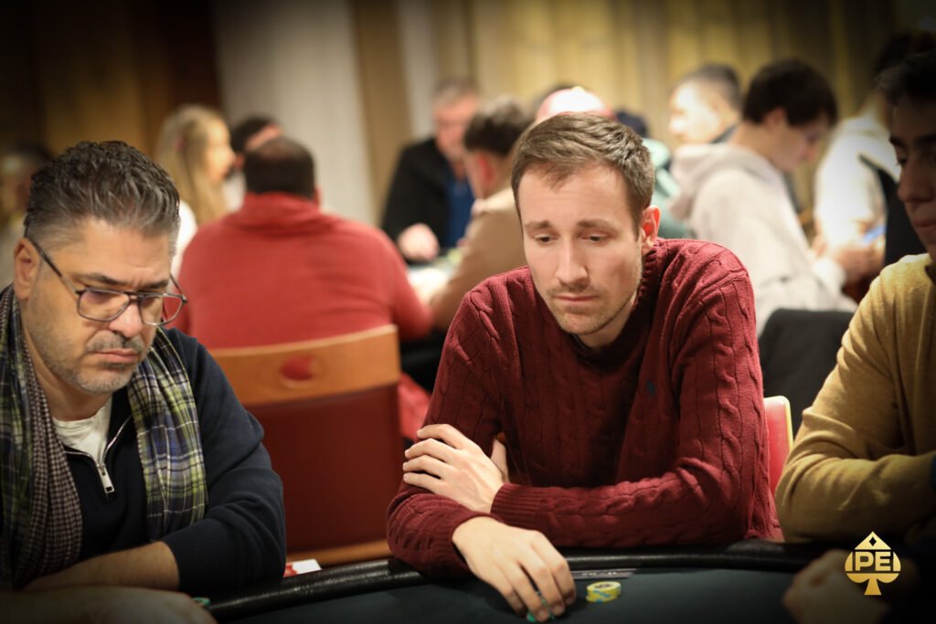 Ein Mann in einem schwarzen Anzug und einer schwarzen Sonnenbrille sitzt an einem Pokertisch und hält seine Spielkarten in der Hand. Im Hintergrund sind andere Spieler und Pokerchips zu sehen.
