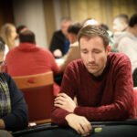 Ein Mann in einem schwarzen Anzug und einer schwarzen Sonnenbrille sitzt an einem Pokertisch und hält seine Spielkarten in der Hand. Im Hintergrund sind andere Spieler und Pokerchips zu sehen.