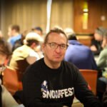 Ein professioneller Pokertisch mit grünem Filz, umgeben von Stühlen und Spielkarten, Chips und Getränken. Zwei Personen sitzen am Tisch und halten ihre Karten, während sie konzentri