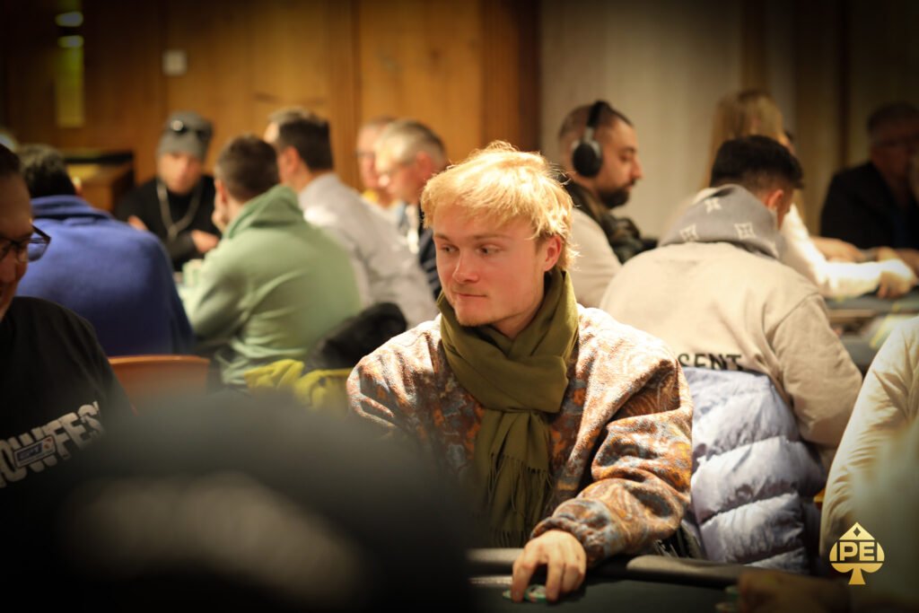 Ein Mann in einem dunkelblauen Anzug und einer weißen Krawatte sitzt an einem Pokertisch und hält zwei Spielkarten in der Hand. Er schaut konzentriert auf den Tisch, auf dem Poker